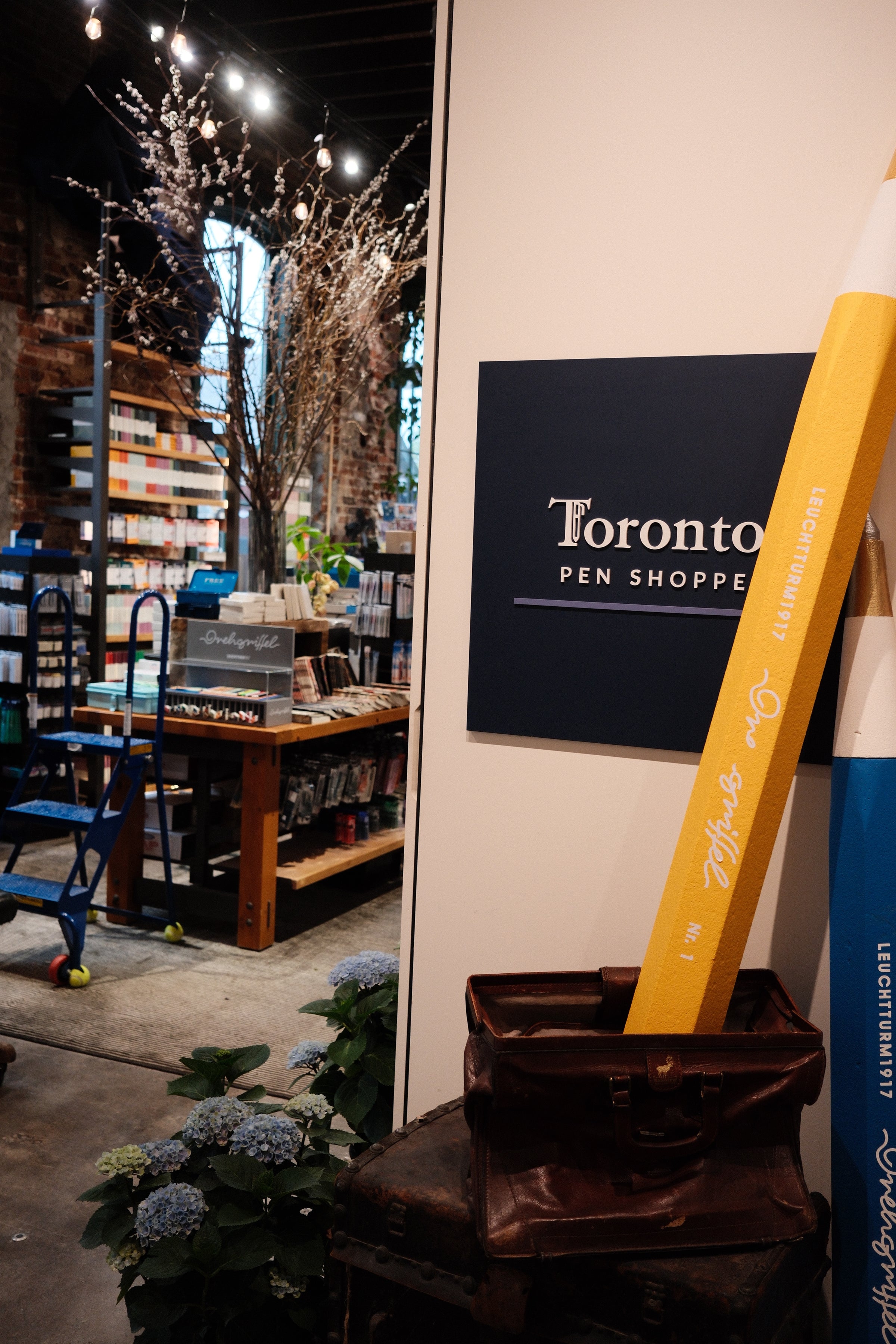Toronto Pen Shoppe sign with a large yellow pen in front, indoor setting with plants and furniture.